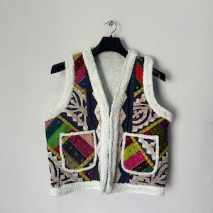 May include: A colorful patchwork vest with a white, fluffy trim and two front pockets. The vest features a variety of patterned fabrics in shades of blue, green, red, and yellow. It is hanging on a black hanger against a white background.