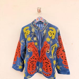 May include: A blue jacket featuring vibrant orange tiger embroidery with black stripes and yellow floral accents. The garment has a stand-up collar, long sleeves, and a blue and white vertical striped lining visible at the open front opening.