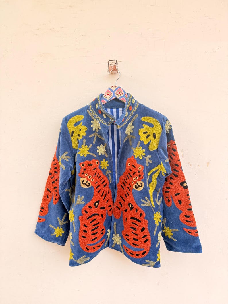 May include: A blue jacket featuring vibrant orange tiger embroidery with black stripes and yellow floral accents. The garment has a stand-up collar, long sleeves, and a blue and white vertical striped lining visible at the open front opening.