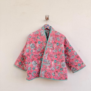 May include: A quilted jacket featuring a vibrant pink base with a repeating floral and botanical print in shades of teal, grey, and white. The garment has three-quarter length sleeves, an open front, and is finished with a green and white striped trim.