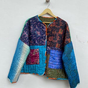 Może przedstawiać: Kolorowa kurtka patchwork z pikowanym wzorem. Kurtka ma mieszankę wzorów i odcieni niebieskiego, fioletowego i pomarańczowego. Wisi na drewnianym wieszaku.