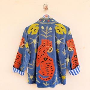 May include: A blue jacket featuring vibrant orange tiger embroidery, yellow leaf patterns, and small floral motifs. The garment has a high collar and long sleeves with cuffs that display a white and blue vertical striped pattern on the interior lining.