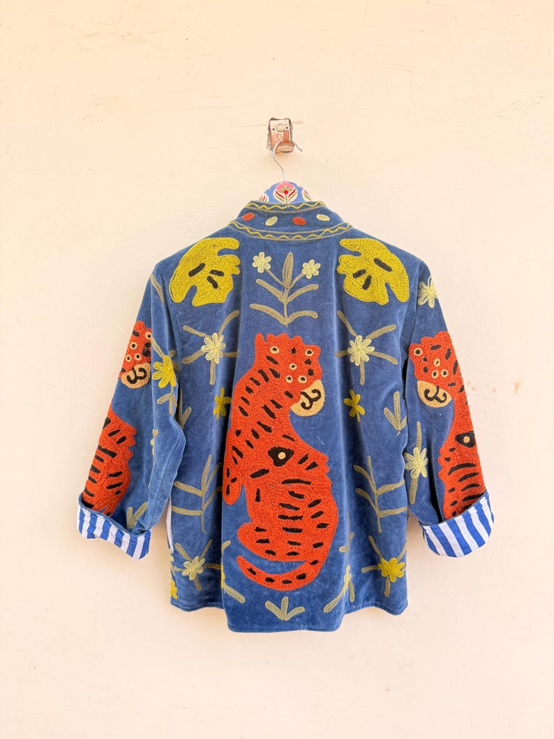 May include: A blue jacket featuring vibrant orange tiger embroidery, yellow leaf patterns, and small floral motifs. The garment has a high collar and long sleeves with cuffs that display a white and blue vertical striped pattern on the interior lining.