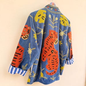 May include: A blue velvet jacket featuring vibrant orange tiger embroidery, yellow leaf motifs, and small floral patterns. The jacket has a high collar and long sleeves with cuffs that display a white and blue vertical striped pattern on the interior.