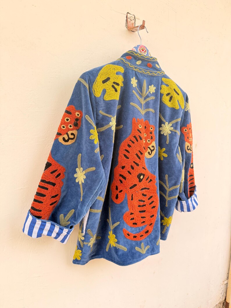 May include: A blue velvet jacket featuring vibrant orange tiger embroidery, yellow leaf motifs, and small floral patterns. The jacket has a high collar and long sleeves with cuffs that display a white and blue vertical striped pattern on the interior.