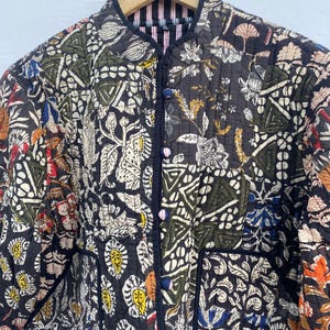 May include: A quilted jacket with a black background and a patchwork design featuring floral and geometric patterns in various colours. The jacket has a mandarin collar, button closure, and black trim. The jacket is hanging on a wooden hanger.
