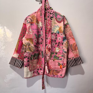 May include: A patchwork kimono-style jacket in shades of pink, featuring floral and geometric patterns. The jacket has a tie closure, pockets, and black and white striped cuffs. It is hanging on a metal hook.