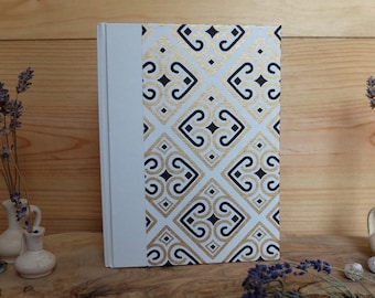 May include: A white hardcover journal with a gold and black geometric pattern on the cover. The journal is sitting on a wooden surface with two small vases of dried lavender.