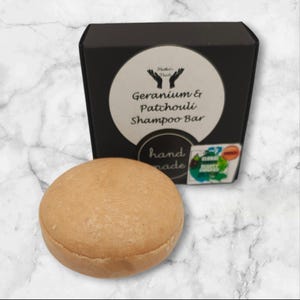 May include: A round, light brown shampoo bar in a black box with the text "Geranium & Patchouli Shampoo Bar" and "hand made".