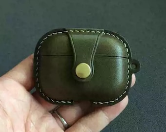 May include: Olive green leather AirPod case with white stitching and a brass button closure. The case is rectangular with rounded edges and a small loop on the side. A stylish accessory for protecting your wireless earbuds.
