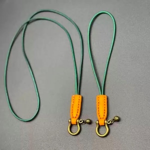 Puede incluir: Dos cordones verdes con detalles de cuero naranja y herrajes de latón antiguo. Cada cordón cuenta con un cierre en forma de U y una pequeña cuenta decorativa. Los cordones están diseñados para sujetar llaves o pequeños accesorios.