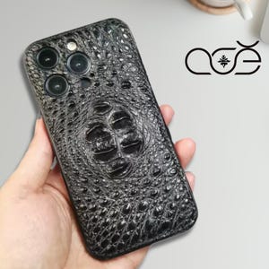 Może przedstawiać: Czarny, teksturowany pokrowiec na telefon z wzorem skóry krokodyla. Etui posiada trzy wycięcia na aparat i wycięcie na lampę błyskową. Logo marki "ase" jest widoczne w prawym górnym rogu.
