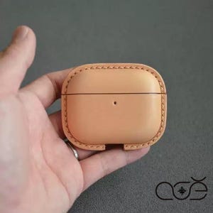 Könnte beinhalten: Eine hellbraune Schutzhülle aus Leder für kabellose Ohrhörer mit einer glatten, abgerundeten rechteckigen Form, sichtbaren handgenähten Kanten entlang des Umfangs und einer kleinen kreisförmigen Aussparung an der Vorderseite für die Ladeanzeige.