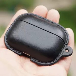 Puede incluir: Una funda de cuero negro para auriculares inalámbricos. La funda es rectangular con bordes redondeados y un borde cosido. Tiene un pequeño lazo para sujetar a un llavero o cordón. La funda se sostiene en una mano.