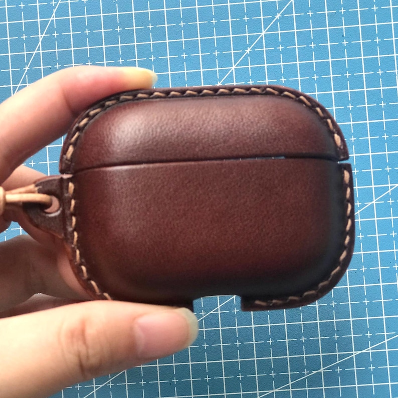 K&ouml;nnte beinhalten: Eine braune Leder-AirPod-H&uuml;lle mit einer gen&auml;hten Umrandung. Die H&uuml;lle hat eine glatte Oberfl&auml;che und eine kleine Schlaufe zum Befestigen eines Schl&uuml;sselbands. Die H&uuml;lle wird in einer Hand vor einem blauen Gitterhintergrund gehalten.