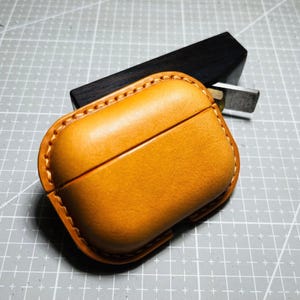 K&ouml;nnte beinhalten: Handgefertigte, hellbraune Leder-AirPods-H&uuml;lle mit gen&auml;htem Rand. Die H&uuml;lle ist ge&ouml;ffnet und gibt den Blick ins Innere frei. Ein dunkler Holzblock und ein Metallwerkzeug befinden sich im Hintergrund auf einer grauen Schneidematte.