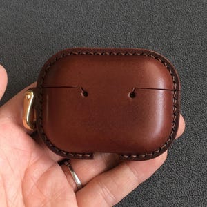 May include: A brown leather AirPod case with a stitched border and a gold-colored clasp. The case is rectangular with rounded edges and two small holes on the top. The leather has a rich, warm tone.