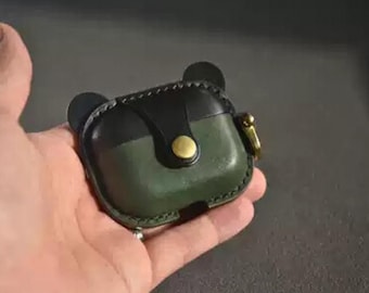 May include: A handcrafted leather AirPod case in a unique bear design. The case features a two-tone color scheme with a black top and olive green base. It has a gold-colored button closure and a small gold-colored clasp.