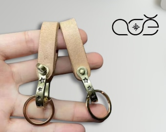 May include: Two leather keychains with brass hardware. Each keychain features a light brown leather strap, a brass clasp with star cutouts, and a brass ring. The keychains are designed for holding keys.