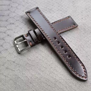 May include: A dark brown leather watch strap with orange stitching. The strap has a silver-coloured buckle and multiple adjustment holes. The leather has a polished finish, and the stitching adds a decorative element.