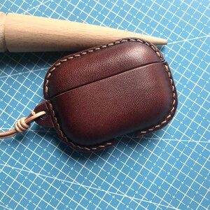 K&ouml;nnte beinhalten: Ein weinrotes Leder-AirPod-Case mit einer gen&auml;hten Bord&uuml;re und einem passenden Lederriemen. Das Case liegt auf einer blauen Schneidematte mit einem Holzd&uuml;bel im Hintergrund.