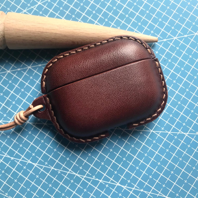 K&ouml;nnte beinhalten: Ein weinrotes Leder-AirPod-Case mit einer gen&auml;hten Bord&uuml;re und einem passenden Lederriemen. Das Case liegt auf einer blauen Schneidematte mit einem Holzd&uuml;bel im Hintergrund.