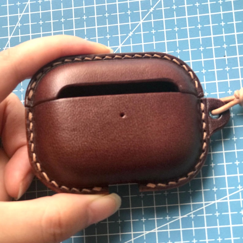 K&ouml;nnte beinhalten: Eine braune Leder-AirPod-H&uuml;lle mit einer gen&auml;hten Bord&uuml;re und einem kleinen Loch. Die H&uuml;lle hat einen Ausschnitt f&uuml;r den Ladeanschluss und eine Schlaufe f&uuml;r ein Schl&uuml;sselband. Die H&uuml;lle wird in einer Hand gehalten.