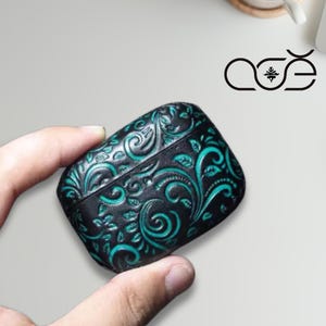 May include: Black and teal leather case with an embossed floral design. The case has a rectangular shape and is held in a hand. The case has a decorative pattern and a logo in the upper right corner.