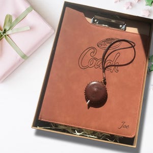 Op de afbeelding: Een bruin lederen klembord met een gouden metalen clip en een bruin lederen band met een metalen ring. Het klembord heeft het woord "Coach" gegraveerd, samen met een voetbalontwerp. De band heeft een kleine ronde hanger met het woord "Coach" gegraveerd. Het klembord zit in een doos met bruin papier als vulling.