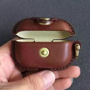 May include: A brown leather AirPod case with a hinged lid, open to reveal a cream-colored interior. The case features brass-colored hardware, including a clasp and a circular button on the front. The case is held open by a hand.