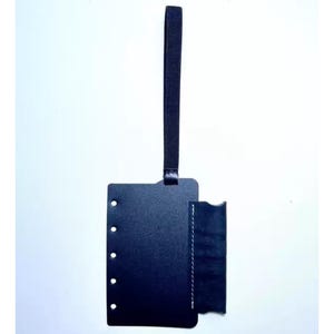 Puede incluir: Una etiqueta de equipaje de cuero negro con un asa de bucle. La etiqueta tiene forma rectangular con esquinas redondeadas y un bolsillo lateral. Hay agujeros a lo largo del borde izquierdo. El bolsillo está cosido con hilo blanco.