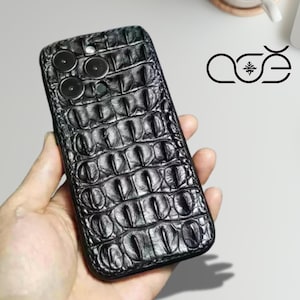Może przedstawiać: Czarny futerał na telefon z fakturą skóry krokodyla. Etui posiada wycięcia na obiektywy aparatu telefonu. Logo marki jest widoczne w prawym górnym rogu. Etui jest trzymane w dłoni.