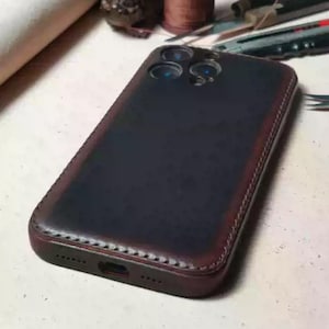Może przedstawiać: Ciemnoniebieskie skórzane etui na telefon z czerwonobrązową obwódką. Etui ma szytą krawędź i wycięcia na aparat i port ładowania telefonu. Etui jest przeznaczone do smartfona.