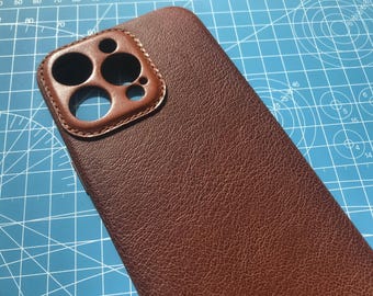 May include: A brown leather phone case with a cutout for the camera lens. The case has a textured surface and a stitched edge.