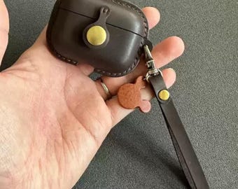 May include: Dark brown leather AirPod case with a gold-colored button closure and matching strap. The case has a stitched border and a small, decorative leather charm. The strap is attached with silver-colored hardware.