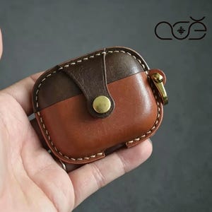 Peut inclure: Un étui de protection bicolore en cuir marron pour écouteurs sans fil, doté d'une fermeture à rabat avec un bouton-pression en métal doré, de surpiqûres blanches et d'un anneau en métal doré sur le côté. Le logo ace est visible.