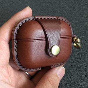 May include: A brown leather AirPod case with a brass button closure and stitched edging. The case features a small leather strap and a brass ring for attachment. The case is held in a hand.