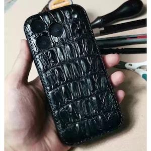 Puede incluir: Funda de teléfono de cuero negro con un patrón de piel de cocodrilo texturizado. La funda tiene un recorte para la cámara y un diseño elegante y ajustado. La funda está diseñada para proteger el teléfono de arañazos e impactos.
