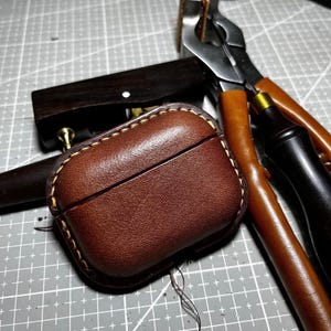 K&ouml;nnte beinhalten: Eine braune Leder-AirPod-H&uuml;lle mit gelben N&auml;hten. Die H&uuml;lle ist rechteckig und liegt auf einer grauen Oberfl&auml;che mit einem Rastermuster. Lederwerkzeuge, darunter eine Zange und ein Holzwerkzeug, befinden sich im Hintergrund.