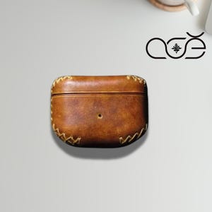 Puede incluir: Una funda de cuero marrón con un diseño cosido, probablemente para auriculares o pequeños aparatos electrónicos. La funda rectangular tiene bordes redondeados y un pequeño agujero central. La costura es de un color más claro que la funda.