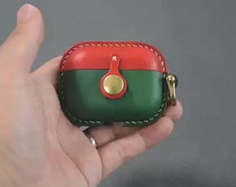 May include: A red and green leather AirPod case with a gold-colored clasp. The case is stitched with white thread and has a gold-colored button closure. The case is held in a hand.