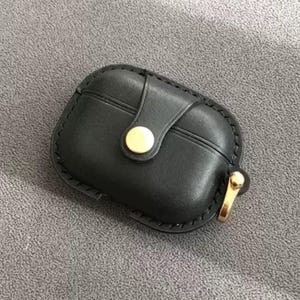 Peut inclure: Étui noir en cuir pour AirPod avec fermeture à bouton doré et boucle en métal doré. L'étui a une bordure cousue et un rabat qui se ferme avec un bouton-pression. Conçu pour protéger et transporter des écouteurs sans fil.