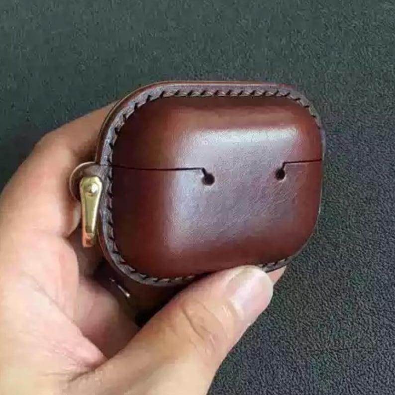 May include: A brown leather AirPod case with a stitched border and gold-colored clasp. The case is rectangular with rounded edges and two small holes on the front. The case is held in a hand.