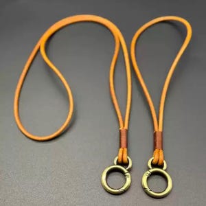 May include: A tan leather lanyard with two bronze-colored clasps. The lanyard is made of a thin, round leather cord. The clasps are circular with a hinged opening. The lanyard is designed to be worn around the neck.