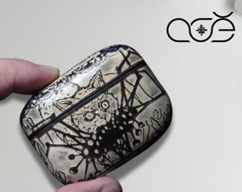 May include: A decorative, rectangular case with a detailed, black and white illustration. The case features a stylized design with a central creature and floral elements. The case is held in a hand. The logo "aöe" is visible in the background.