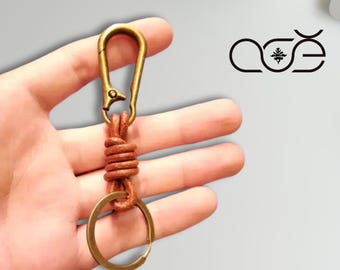 May include: A brown leather keychain with a brass-colored clasp and key ring. The leather is wrapped around the connecting hardware. The keychain is held in a hand.