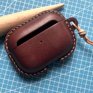 K&ouml;nnte beinhalten: Eine dunkelbraune Leder-AirPod-H&uuml;lle mit einer gen&auml;hten Bord&uuml;re und einer Lederschnur. Die H&uuml;lle ist ge&ouml;ffnet und gibt den Blick ins Innere frei. Der Hintergrund ist eine blaue Schneidematte mit wei&szlig;en Gitterlinien.