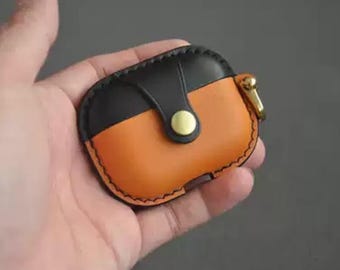 May include: A two-tone leather AirPod case in black and orange. The case has a snap closure with a gold-coloured button and a gold-coloured metal clasp. The case is held in a hand.