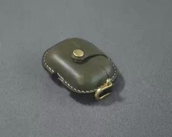 May include: Olive green leather AirPod case with a brass clasp and button closure. The case has white stitching around the edges and a small loop for attaching to a keychain. A stylish accessory for protecting wireless earbuds.