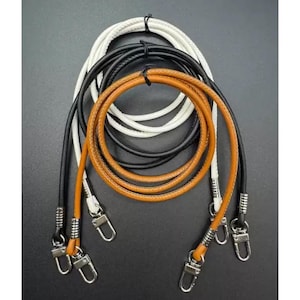 May include: A collection of three leather bag straps in white, black, and tan. Each strap has a silver-toned metal clasp and is designed for easy attachment to a purse or handbag. The straps are made of a smooth, durable leather.